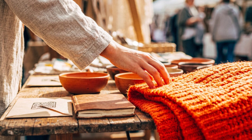 Hand reaches to touch a soft, orange knitted blanket displayed on a rustic wooden table at an outdoor market.